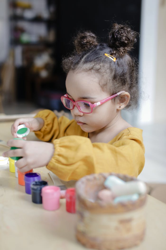 A child wearing glasses paints with colorful containers indoors. Bright and focused moment.