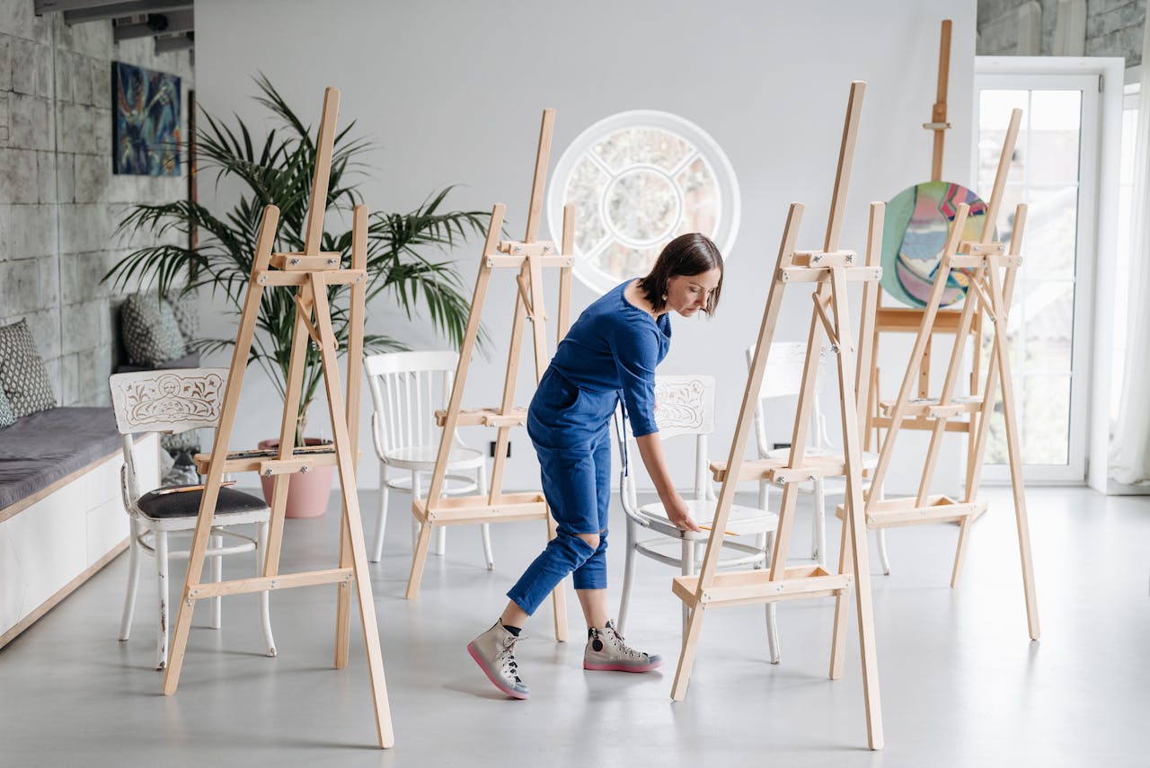 A woman setting up an art studio with wooden easels and white chairs, a creative workspace.