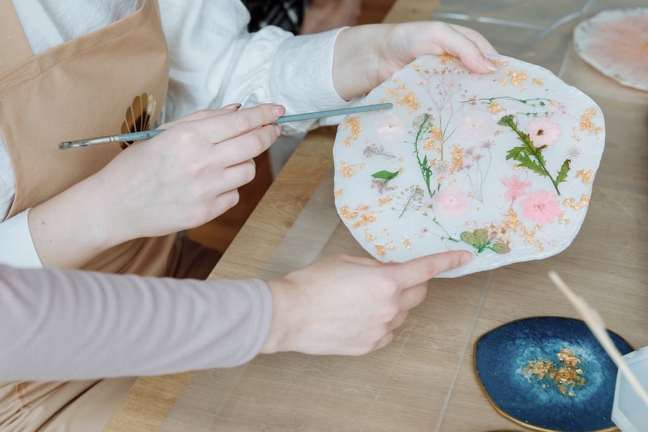 Close-up of hands creating resin artwork with dried flowers and gold flakes in a workshop setting.