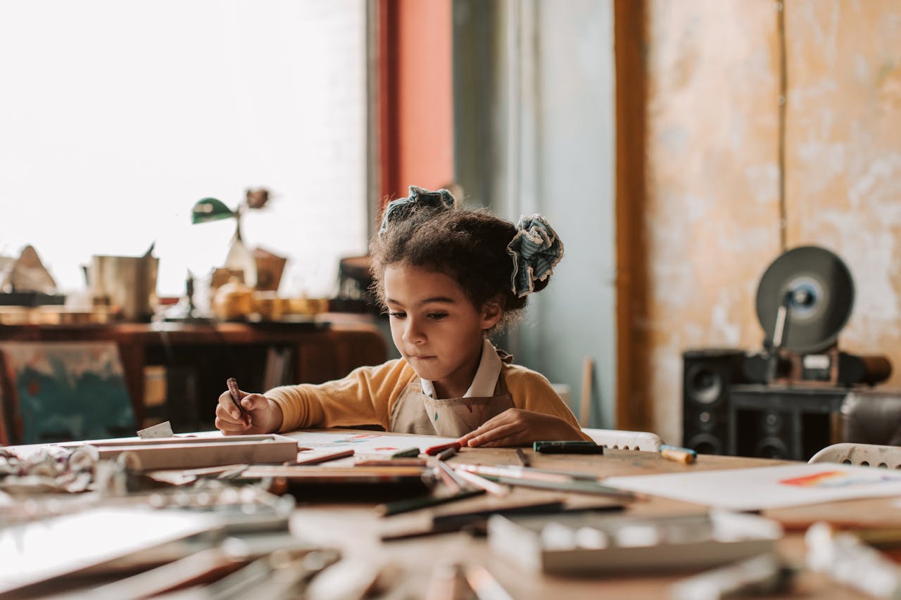 A young girl intensely focused on drawing at a table filled with art supplies, showcasing creativity and art exploration.